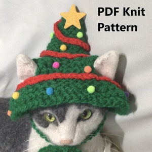 May include: A white cat wearing a green and red knitted Christmas tree hat with a yellow star on top. The hat has colorful pom-poms around the brim.  PDF Knit Pattern.
