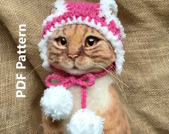 May include: A brown and white cat wearing a pink crocheted hat with white pom poms and a matching pink scarf with white pom poms.