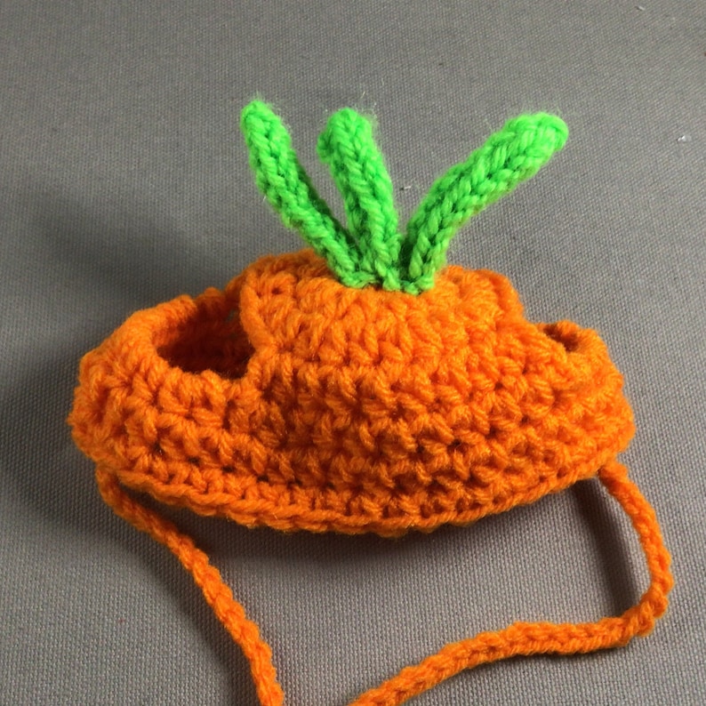May include: Orange crocheted carrot hat with green crocheted leaves on top. The hat has a strap that ties under the chin.