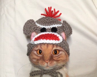 May include: A brown and gray knitted monkey hat with a red pom-pom on top. The hat is worn by a cat with orange and white fur.