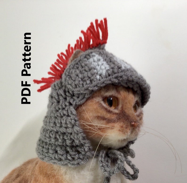 K&ouml;nnte beinhalten: Ein grauer geh&auml;kelter Katzenhelm mit einem roten und wei&szlig;en Kamm. Der Helm hat einen Kinnriemen und wird von einer Katze getragen.