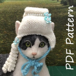 May include: White knitted cat hat with a blue crocheted bow and blue flower embellishments. The hat has a long white crocheted tail.