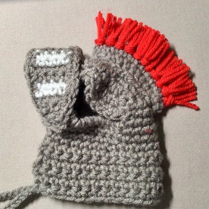 K&ouml;nnte beinhalten: Grauer H&auml;kelhelm mit roter Fransenborte oben. Der Helm hat ein wei&szlig;es H&auml;kelmuster an der Seite.