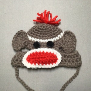 May include: A brown crocheted monkey hat with red yarn hair, white and red crocheted mouth, and black button eyes.