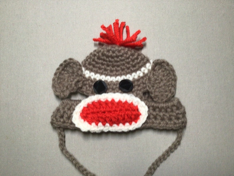 May include: A brown crocheted monkey hat with red yarn hair, white and red crocheted mouth, and black button eyes.
