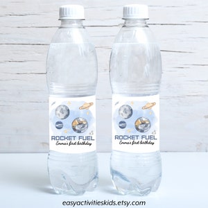 May include: Two clear plastic water bottles with blue and white labels that say "Rocket Fuel Emma's first birthday". The labels feature a watercolor illustration of planets and stars.