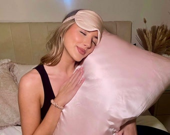 May include: A light pink satin pillow and matching eye mask. The pillow is large and square, with a smooth, shiny surface. The eye mask has a thin elastic band. The items are likely for sleep or relaxation.