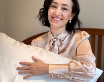 May include: A woman in a light brown satin blouse holds a white satin pillow. The pillow is smooth and has a slight sheen. The woman is smiling and has dark hair.