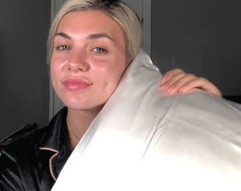 May include: A woman with blonde hair is holding a white pillow in front of her face. She is wearing a black satin shirt with white piping.
