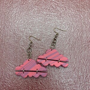 Peut inclure: Une paire de boucles d'oreilles en forme de nuage rose et rouge avec une chaîne et un crochet argentés. Les boucles d'oreilles sont en pâte polymère et ont un motif rayé.