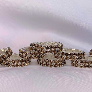 May include: Six brown rhinestone napkin rings, each with a silver metal base. The rings are stacked on top of each other.