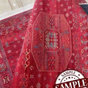 May include: Red rug with a geometric pattern and a white fringe border. The rug has a sample label with the word "SAMPLE" repeated three times.