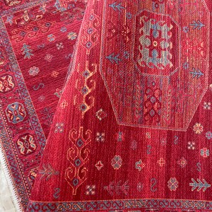 May include: A red rug with a geometric pattern in shades of blue, yellow, and cream. The rug has a fringe border.