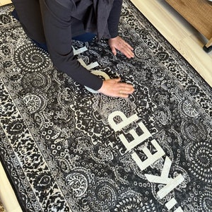 May include: A rectangular area rug featuring a black and white intricate paisley and floral pattern. The rug displays the word KEEP in large, bold, white capital letters oriented vertically across the center of the detailed decorative design.