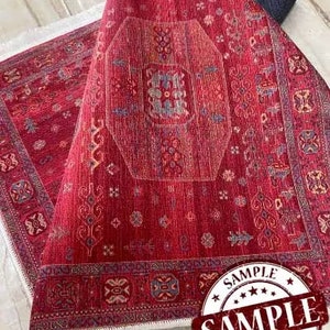 May include: Red rug with a white border and intricate floral patterns. The rug has a central medallion with a geometric design. The rug is labeled "SAMPLE" in a red circle.