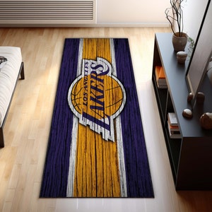 May include: A rectangular rug featuring the Los Angeles Lakers logo. The rug has a wood grain design with purple and gold colors. The Lakers logo is prominently displayed in the center.
