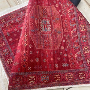 May include: Red rug with a detailed geometric pattern in shades of red, orange, blue, and white. The rug has a white fringe border.