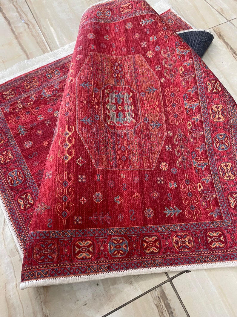 May include: Red rug with a detailed geometric pattern in shades of red, orange, blue, and white. The rug has a white fringe border.