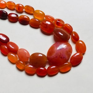 May include: A strand of carnelian gemstone beads in shades of orange and red. The beads are oval shaped and have a smooth, polished finish.