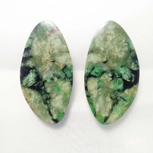 Könnte beinhalten: Zwei ovale Edelsteine mit marmoriertem Aussehen. Die Steine weisen eine Mischung aus grünen, schwarzen und weißen Farbtönen mit polierter Oberfläche auf. Die Edelsteine werden vor einem weißen Hintergrund präsentiert, um ihre natürlichen Muster und Farben hervorzuheben.