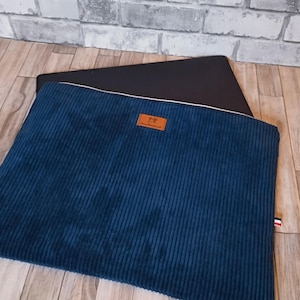 Peut inclure: Une housse pour ordinateur portable en velours côtelé bleu avec un patch en cuir marron et des coutures blanches. La housse est posée sur une surface en bois.