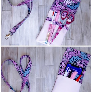May include: A fabric pouch with a floral pattern in shades of purple, pink, and blue, featuring a light pink pocket and a matching lanyard. The pouch displays the text "Aide Soignante" and holds various pens, scissors, or a smartphone.