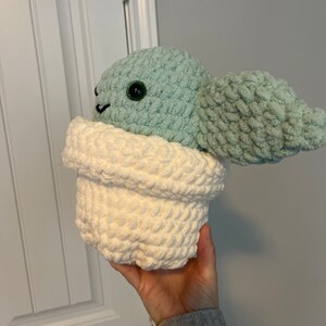 May include: A crocheted plush toy in the shape of a green character with a white pot. The character has large green eyes and a small mouth.