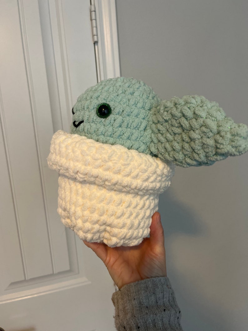 May include: A crocheted plush toy in the shape of a green character with a white pot. The character has large green eyes and a small mouth.