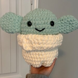 May include: A crocheted plush toy of a green alien character with big eyes and a white crocheted scarf. The toy is being held by a hand.