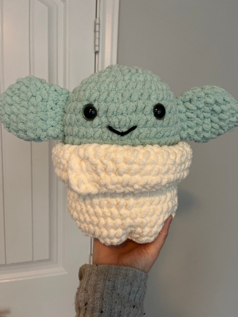 May include: A crocheted plush toy of a green alien character with big eyes and a white crocheted scarf. The toy is being held by a hand.