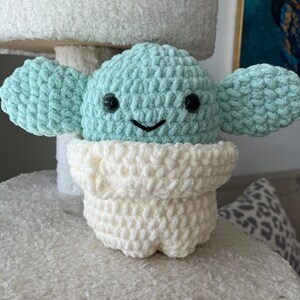 May include: A crocheted plush toy in the shape of a green alien character with big black eyes and a white crocheted robe. The toy is sitting on a white fuzzy surface.