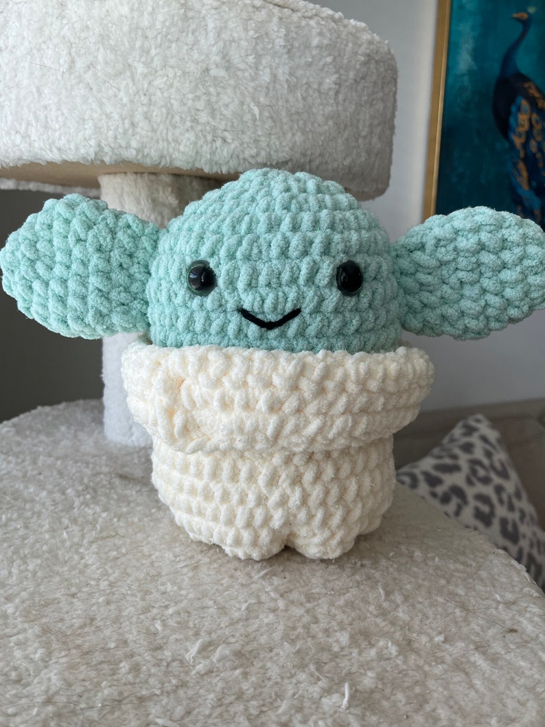 May include: A crocheted plush toy in the shape of a green alien character with big black eyes and a white crocheted robe. The toy is sitting on a white fuzzy surface.