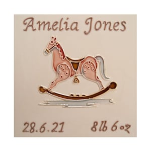 May include: A personalized ceramic tile with a pink rocking horse illustration, the name "Amelia Jones", the date "28.6.21", and the weight "8 lb 6 oz".