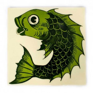 May include: Green ceramic tile with a stylized image of a fish. The fish has a large eye and a wide open mouth. The tile has a white background.