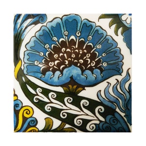 Puede incluir: Azulejo cerámico cuadrado con un diseño floral detallado. La flor central es de un azul vibrante con un centro marrón oscuro y detalles blancos. Hojas y tallos verdes con volutas blancas completan el diseño, sobre un fondo blanco.