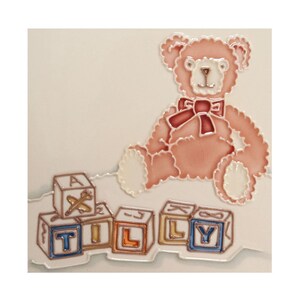 May include: A ceramic tile with a pink teddy bear wearing a red bow and holding a set of colorful alphabet blocks that spell "TILLY".
