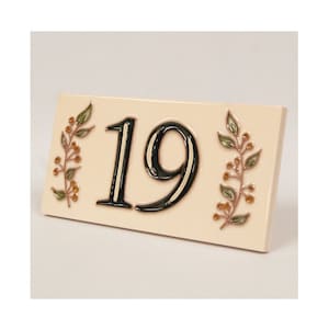 May include: A ceramic house number plaque with the number 19 in black with a white outline. The plaque is cream colored with a floral design in green and brown.