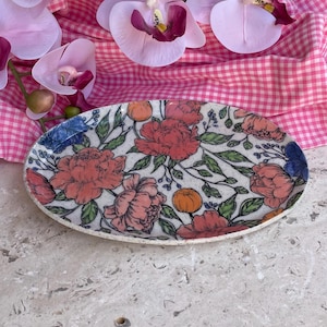 May include: Oval ceramic serving platter with a floral design. The platter features a white background with red, orange, and blue flowers and green leaves. The edges are slightly raised and the platter is approximately 30 cm long.