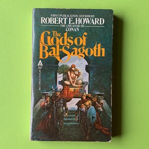 Könnte beinhalten: Vintage Taschenbuch "The Gods of Bal-Sagoth" von Robert E. Howard. Das Cover zeigt eine Fantasy-Illustration mit einer sitzenden Frau und Kriegern. Das Buch hat ein grünes und dunkelblaues Cover mit dem Titel und dem Namen des Autors.