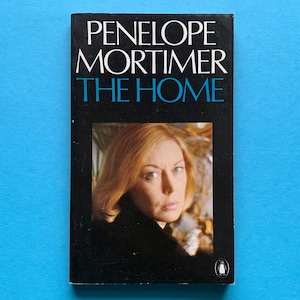 May include: A vintage paperback book titled "THE HOME" by Penelope Mortimer. The cover is black with white and teal lettering. A portrait of a woman with blonde hair is featured in a framed image on the cover. The book is set against a bright blue background.