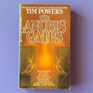 May include: A vintage paperback book titled "The Anubis Gates" by Tim Powers. The cover is predominantly brown with gold lettering and features an illustration of two Anubis figures. The text on the cover reads "Backward in time to solve an ancient mystery...and create some new ones."