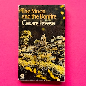 May include: A vintage paperback book, "The Moon and the Bonfire" by Cesare Pavese. The cover shows a village scene in yellow and white against a dark background. The book is set against a bright pink backdrop. The publisher is Quarter.