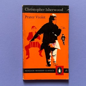 May include: Vintage paperback book titled "Prater Violet" by Christopher Isherwood. The cover features a striking orange background with black and red illustrations of figures. The Penguin Modern Classics logo is visible.