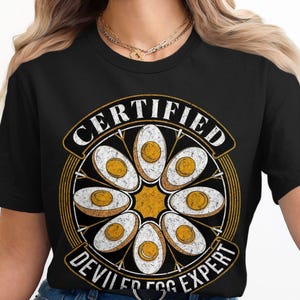 Può includere: Maglietta nera con una grafica bianca di un cerchio di uova ripiene con il testo "CERTIFIED DEVILLED EGG EXPERT".