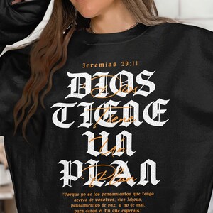 May include: Black sweatshirt featuring the Spanish text "DIOS TIENE UN PLAN" in white gothic font, with the bible verse "Jeremias 29:11". The sweatshirt also has a quote in Spanish. A casual, faith-based apparel item.