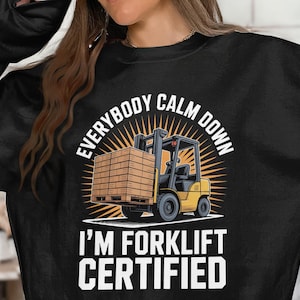 May include: Black sweatshirt with a graphic of a yellow forklift carrying a pallet of wood. The text reads "EVERYBODY CALM DOWN I'M FORKLIFT CERTIFIED" in white block letters and a white sunburst design.