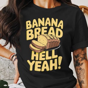 Può includere: Maglietta nera con una grafica gialla che dice "Banana Bread Hell Yeah!" e un'immagine di un pane al banana con fette.
