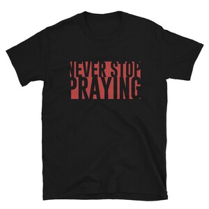 Puede incluir: Camiseta negra de manga corta con el texto "NEVER STOP PRAYING" en rojo.