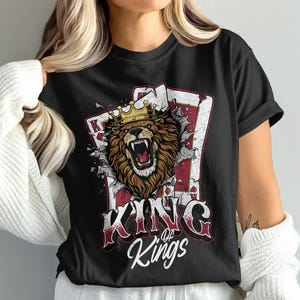 以下が含まれることがあります： 金色の王冠をかぶったライオンがトランプを突き破る黒いTシャツ。「KING of Kings」の文字はかすれたフォントで表示されています。大胆で人目を引くデザインです。Tシャツは定番のクルーネックです。