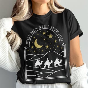 May include: Black t-shirt with a white graphic of three camels, a crescent moon, stars, and the text "WISE MEN STILL SEEK HIM." The design is framed by a white archway, evoking a desert night scene.